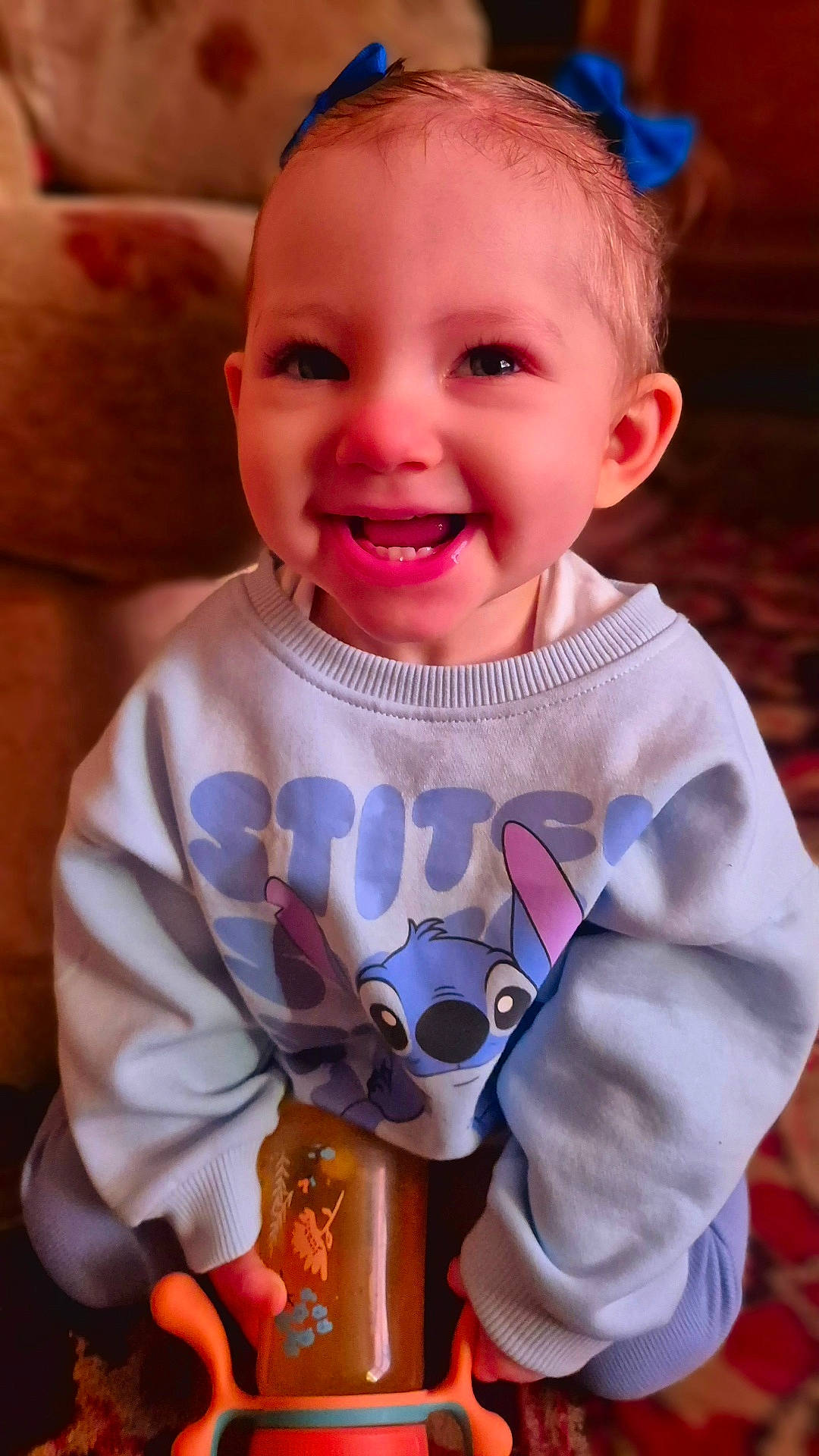 baby, child, clothing, cute, expression, face, fun, happy, indoor, joy, person, sippy_cup, sitting, sitting_on_floor, smiling, stitch, sweatshirt, toddler, toy, warm