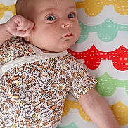 Romy participe au concours pour gagner de l'argent avec cette photo : baby, infant, child, floral_clothing, patterned_mat, colorful_background, lying_down, fist, curious_eyes, cute, indoor, soft_lighting, skin, head, arm, onesie, person, small_child, baby_garment, playmat