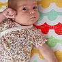 baby, infant, child, floral_clothing, patterned_mat, colorful_background, lying_down, fist, curious_eyes, cute, indoor, soft_lighting, skin, head, arm, onesie, person, small_child, baby_garment, playmat