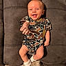 baby, smiling, camo_onesie, white_socks, couch, gray_furniture, infant, happy, reaching_hand, indoor, person, child, cute, plush, seated, young_baby, cozy, portrait, casual_clothing, relaxed