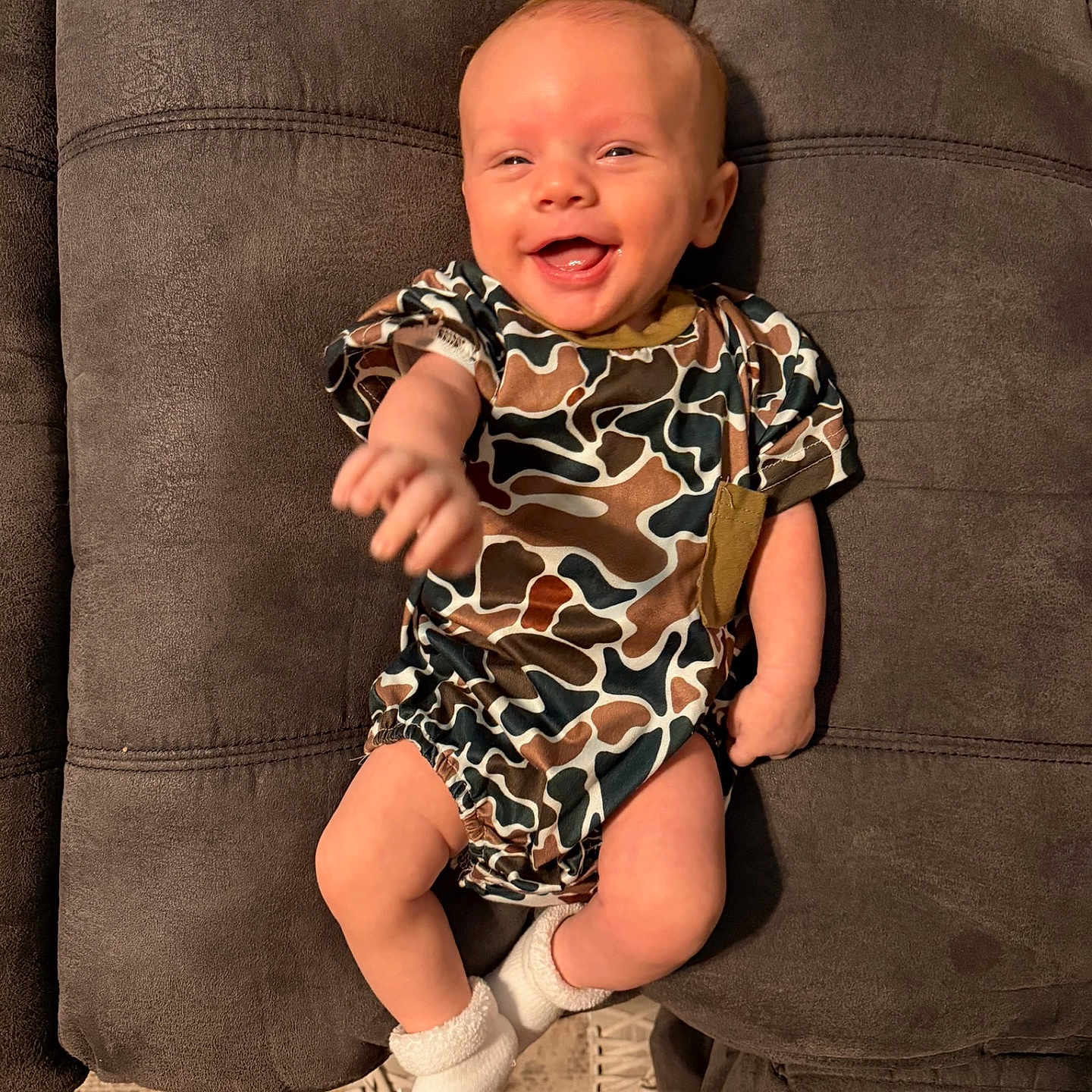 Crue joined the competition — help win amazing prizes! baby, smiling, camo_onesie, white_socks, couch, gray_furniture, infant, happy, reaching_hand, indoor, person, child, cute, plush, seated, young_baby, cozy, portrait, casual_clothing, relaxed