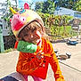 Rosie is registered to the contest to win money with this photo: child, helmet, unicorn_helmet, toy, water_gun, orange_shirt, pink_pants, outdoor, backyard, sunny, smile, playful, concrete, fence, greenery, chair, toy_vehicle, happy, person, daylight