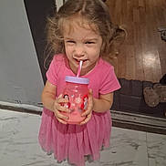 Raven is registered to the contest to win money with this photo: child, girl, pink_dress, drinking, cup, straw, indoor, floor, marble_floor, wooden_floor, door, smiling, happy, person, young_child, curly_hair, playful, drinkware, cartoon_characters, home