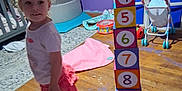 Raven is registered to the contest to win money with this photo: child, toddler, indoor, wooden_floor, stacked_blocks, numbered_blocks, toys, purple_wall, curly_hair, barefoot, skirt, short_sleeve_shirt, crib, toy_stroller, stuffed_animal, rug, storage_bins, playroom, floor_mat, childrens_room