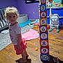 child, toddler, indoor, wooden_floor, stacked_blocks, numbered_blocks, toys, purple_wall, curly_hair, barefoot, skirt, short_sleeve_shirt, crib, toy_stroller, stuffed_animal, rug, storage_bins, playroom, floor_mat, childrens_room