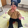 toddler, child, sitting, hat, pom_poms, sweater, black_pants, white_sneakers, finger_in_mouth, play_mat, indoor, floor, radiator, door, curious, cute, baby, person, clothing, expression