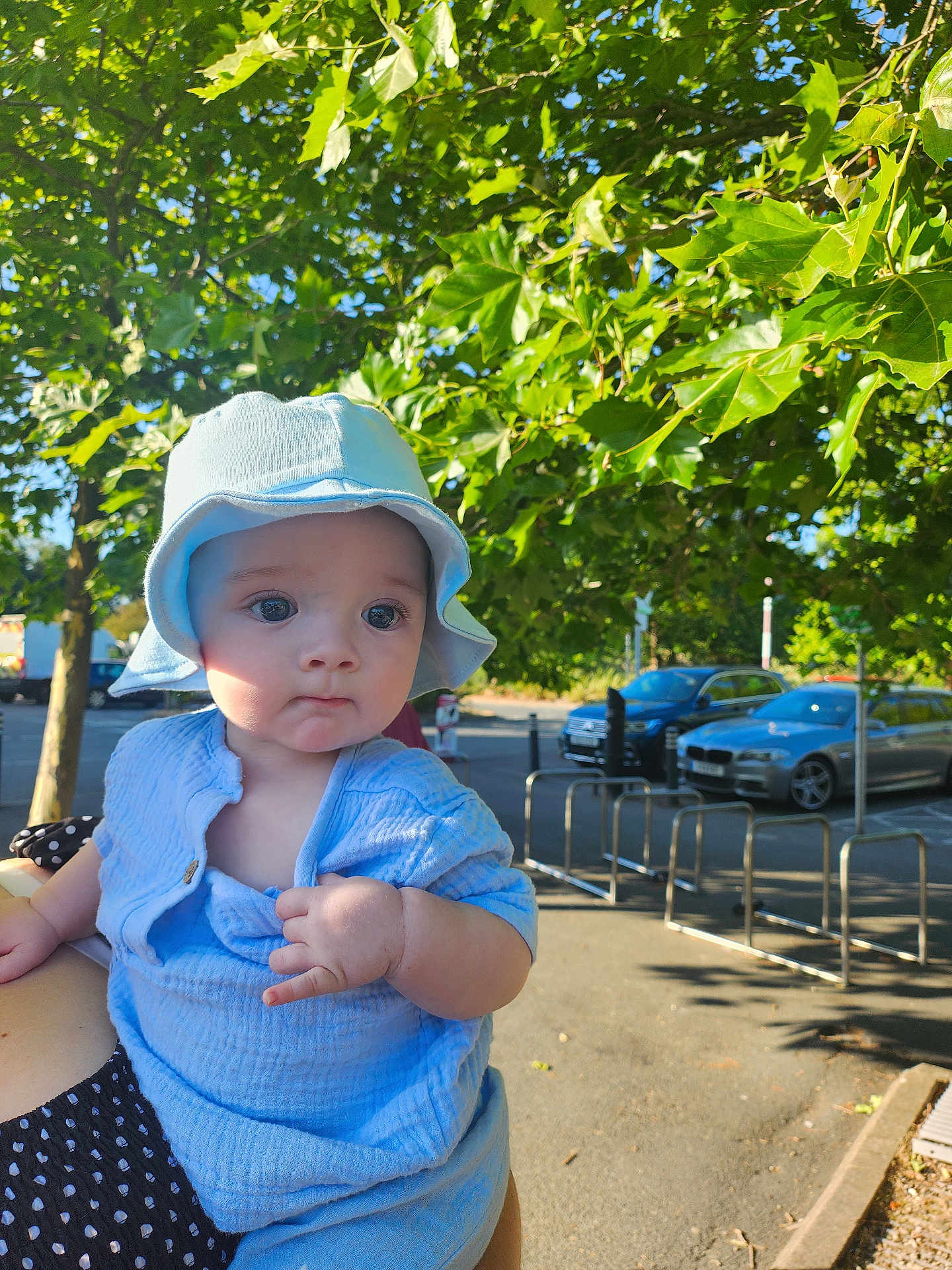 Alex is registered to the contest to win money with this photo: baby, child, blue_clothing, hat, outdoor, sunlight, green_leaves, parking_lot, car, person, holding, facial_expression, cute, summer, nature, daytime, infant, casual, portrait, hand