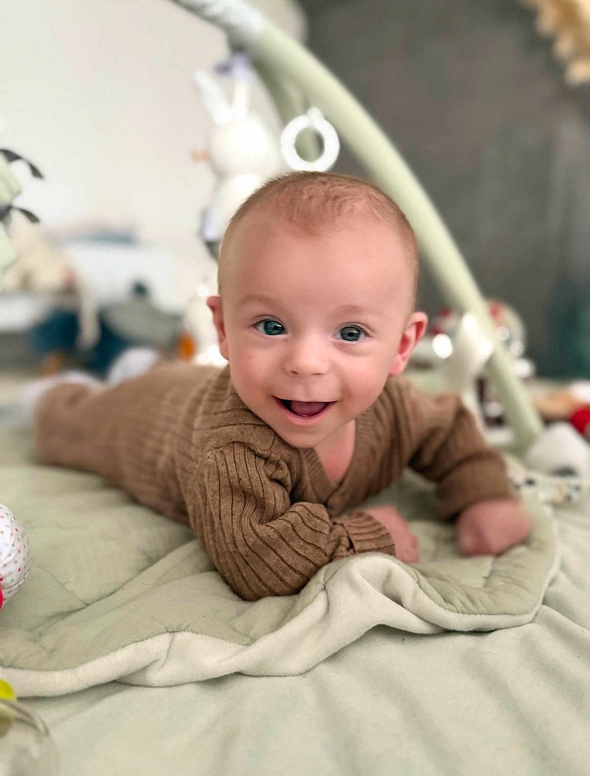 Noé participe au concours pour gagner de l'argent avec cette photo : baby, infant, smiling, face, sweater, brown_clothing, blanket, quilt, lying_down, happy, indoor, soft_toys, child, cute, portrait, head, blue_eyes, skin, person, cozy