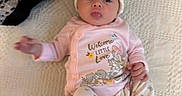 Delaney is registered to the contest to win money with this photo: baby, infant, child, portrait, lying_down, blanket, knitted_blanket, pink_outfit, baby_hat, bunny_hat, clothing, smiling, wide_eyes, indoor, cozy, bed, text_on_clothing, bunny, floral_pattern, feet