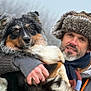 man, dog, winter_clothing, fur_hat, scarf, outdoor, cold_weather, pet, animal, portrait, holding, furry, beard, brown_eyes, black_fur, white_fur, orange_fur, nature, friendship, closeup