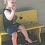 Declan is registered to the contest to win money with this photo: toddler, child, bench, yellow, barefoot, outdoor, sunlight, wooden_floor, heart_cutout, bees, black_shorts, black_shirt, short_hair, playful, sitting, smiling, person, daylight, casual, cute