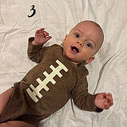 Grayson is registered to the contest to win money with this photo: baby, bed, bedding, brown_outfit, child, eyes, face, hand, happy, indoor, infant, lying_down, number, onesie, open_mouth, playful, portrait, smiling, waving, white_sheet