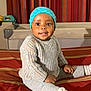 baby, child, smiling, knitted_outfit, blue_hat, bed, striped_bedspread, indoor, crib, cap, person, cute, happy, sitting, cozy, portrait, infant, clothing, face, home
