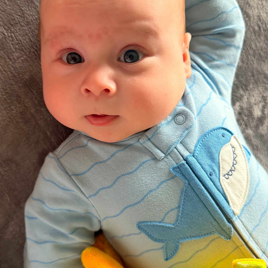 Lucas joined the competition — help win amazing prizes! baby, infant, blue_onesie, plush_toy, yellow_toy, shark_design, blanket, lying_down, cute, child, soft_texture, indoors, portrait, expression, small_hands, face, looking_up, adorable, comfort, casual_clothing