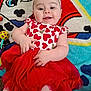child, baby, smiling, bow, heart_pattern, red_skirt, blanket, toy, rattle, carpet, playful, lying_down, portrait, chubby_cheeks, infant, clothing, white_headband, happy, gaze, indoor