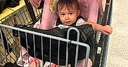 Leyla joined the competition — help win amazing prizes! baby, child, infant, shopping_cart, supermarket, grocery_store, pink_car_seat, safety_harness, seated, portrait, face, eyes, hair, smiling, indoor, produce, fruit, shopping, shopping_aisle, cart