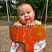 Lilly joined the competition — help win amazing prizes! baby, child, infant, swing, playground, park, outdoor, portrait, smiling, happy, face, hands, blanket, yellow_clothing, orange_swing, rusty_metal, trees, nature, seated, close_up