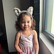 Leslie is registered to the contest to win money with this photo: child, girl, toddler, portrait, smiling, indoors, window, sunlight, dress, pink, cat_ears, headband, curly_hair, brown_hair, standing, wristband, fashion, cute, natural_light, home