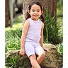Leslie joined the competition — help win amazing prizes! child, girl, smiling, portrait, outdoor, garden, nature, rock, lavender_outfit, pink_clogs, seated, curly_hair, hair_bow, young_child, full_body, tree, plants, sunlight, photograph, screenshot