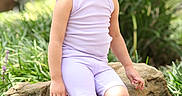 Leslie is registered to the contest to win money with this photo: child, toddler, girl, portrait, sitting, outdoors, garden, rock, tree, plants, grass, nature, lavender_clothing, pink_shoes, clogs, hair_bow, ponytail, looking_up, daylight, smiling