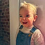 toddler, child, smiling, blue_eyes, blonde_hair, denim_overalls, sweater, portrait, indoor, brick_wall, happy, cute, young_child, face, person, clothing, fashion, casual, expression, light