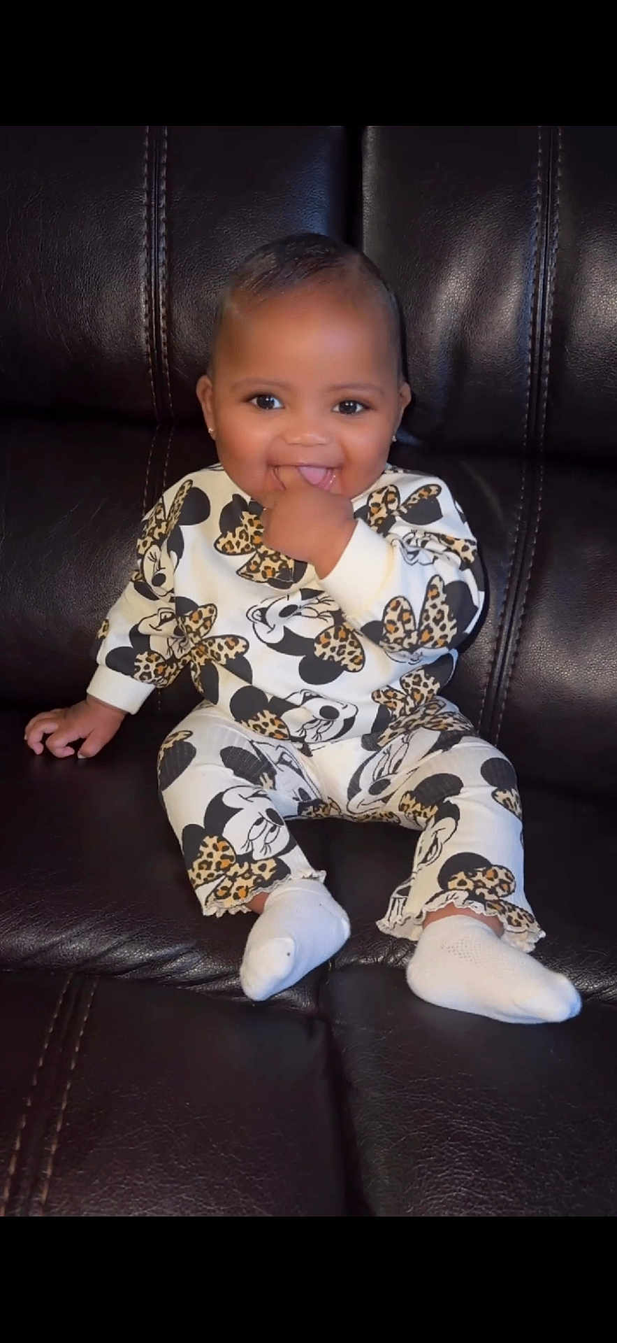 Diyani joined the competition — help win amazing prizes! baby, child, smiling, sitting, couch, leather, outfit, minnie_mouse, patterned_clothing, white_socks, happy, cute, indoor, portrait, young_child, hand_in_mouth, dark_background, small_person, fashion, cheerful