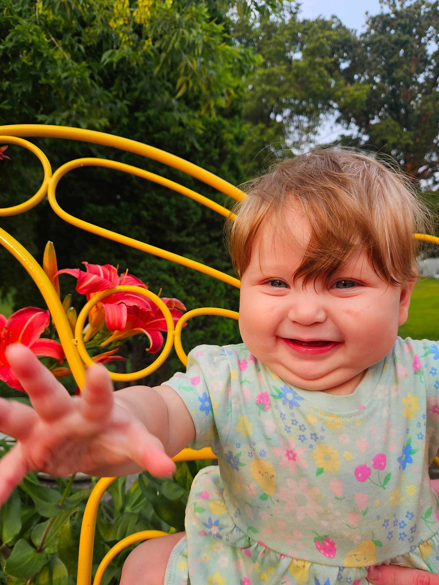 Lillith is registered to the contest to win money with this photo: baby, child, smiling, reaching_hand, yellow_chair, flowers, red_flowers, greenery, outdoor, nature, grass, happy, cute, portrait, toddler, dress, short_sleeves, blond_hair, face, closeup