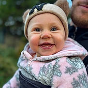 Nicole is registered to the contest to win money with this photo: baby, child, smile, happy, face, hat, beanie, ears, jacket, fleece, pink, gray, pattern, leaf, person, adult, beard, outdoor, blurred_background, closeup