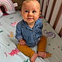 baby, child, infant, crib, bed, bedding, dinosaur, pattern, blue_shirt, mustard_pants, barefoot, cute, sitting, indoors, portrait, smiling, young_child, looking_up, colorful, soft_lighting