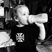 Khaleesi is registered to the contest to win money with this photo: child, toddler, baby, drinking, bottle, milk_bottle, portrait, black_and_white, indoors, home, kitchen, table, chair, shirt, text_on_clothing, ponytail, profile, hand, furniture, family_scene