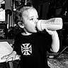 child, toddler, baby, drinking, bottle, milk_bottle, portrait, black_and_white, indoors, home, kitchen, table, chair, shirt, text_on_clothing, ponytail, profile, hand, furniture, family_scene