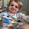 Khaleesi is registered to the contest to win money with this photo: child, toddler, smiling, portrait, indoors, pajamas, cartoon, glasses, toy_story, colorful, sitting, couch, home, wall_art, blonde_hair, face, happy, playful, adult, tattoo