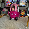 toddler, child, smiling, toy_car, pink, playroom, indoor, television, racing_game, porsche, stuffed_animals, toys, carpet, furniture, speaker, dashboard, sitting, happy, portrait, home