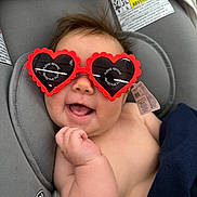 Lyla is registered to the contest to win money with this photo: baby, sunglasses, heart_shaped, car_seat, smiling, infant, protective_film, cheeky, happy, child, clothing_tag, gray, cute, funny, casual, portrait, indoor, head, hand, seat