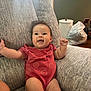 baby, child, smiling, red_onesie, couch, gray_couch, indoor, happy, person, arm_raised, face, home, container, plastic_bag, furniture, sitting, infant, cute, expression, relaxed