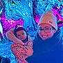 baby, woman, glasses, knitted_hat, scarf, winter_clothing, outdoor, night, colorful_lights, tree_sculpture, face_art, smile, holding, portrait, child, person, forest, decoration, festival, family