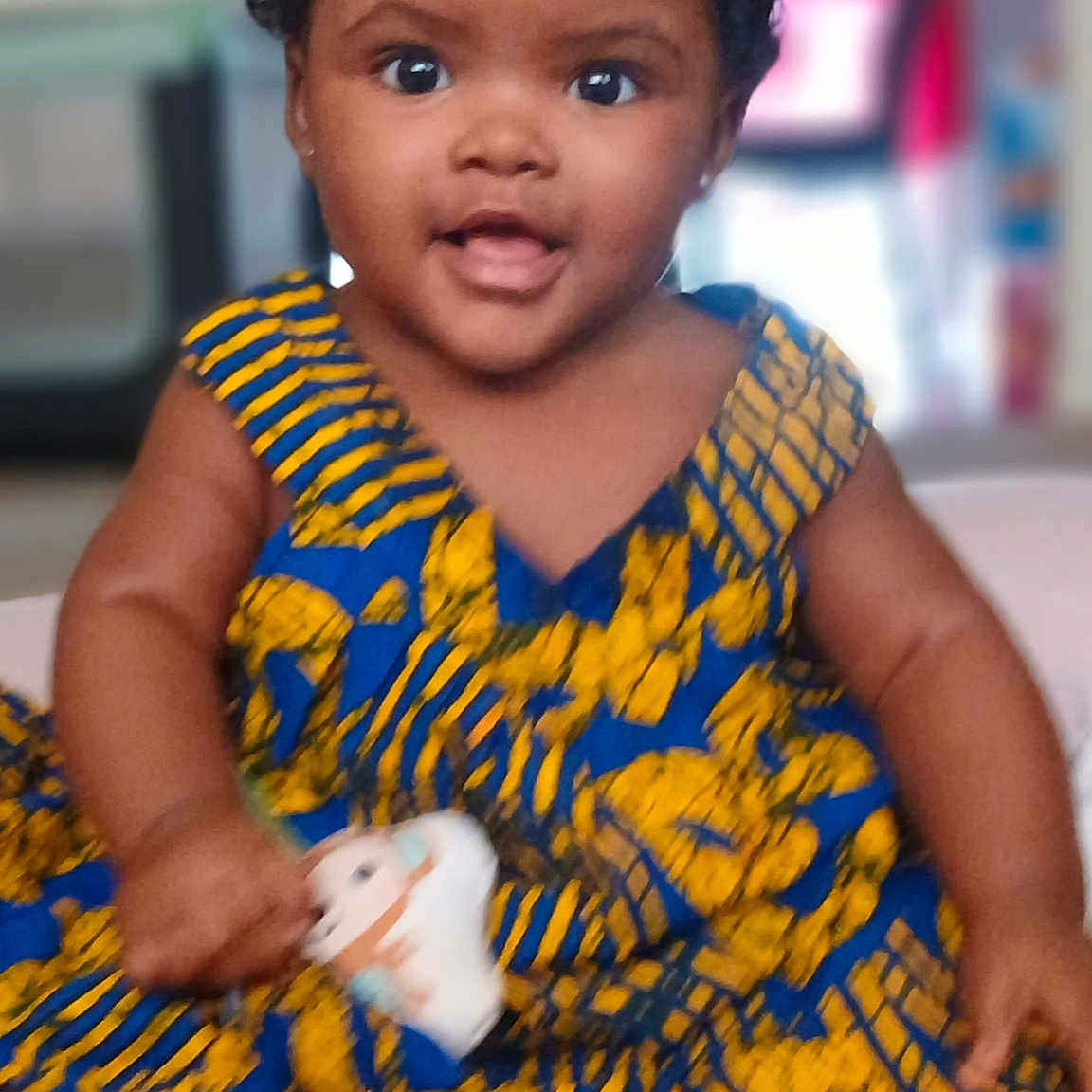 Aminata is registered to the contest to win money with this photo: baby, child, dress, patterned_clothing, toy, indoor, smiling, curly_hair, dark_skin, sitting, happy, infant, portrait, cute, colorful, young_child, barefoot, playful, bright_colors, closeup