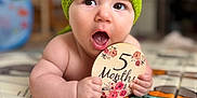 Cléo a rejoint le concours — aidez-le/la à gagner de superbes lots ! baby, infant, green_hat, play_mat, wooden_sign, 5_months, cute, child, indoor, portrait, expression, hand, face, skin, eyes, mouth_open, knitted, floor, colorful, young