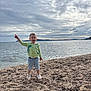 child, toddler, beach, sea, ocean, cloudy_sky, sand, pebbles, water, shore, dinosaur_hoodie, shorts, pointing, standing, smiling, daylight, outdoor, nature, happy, casual_clothing