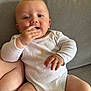 baby, child, infant, onesie, white_clothing, couch, sofa, hand, face, skin, chewing, sitting, indoors, cute, expression, messy_clothing, furniture, comfort, young, portrait