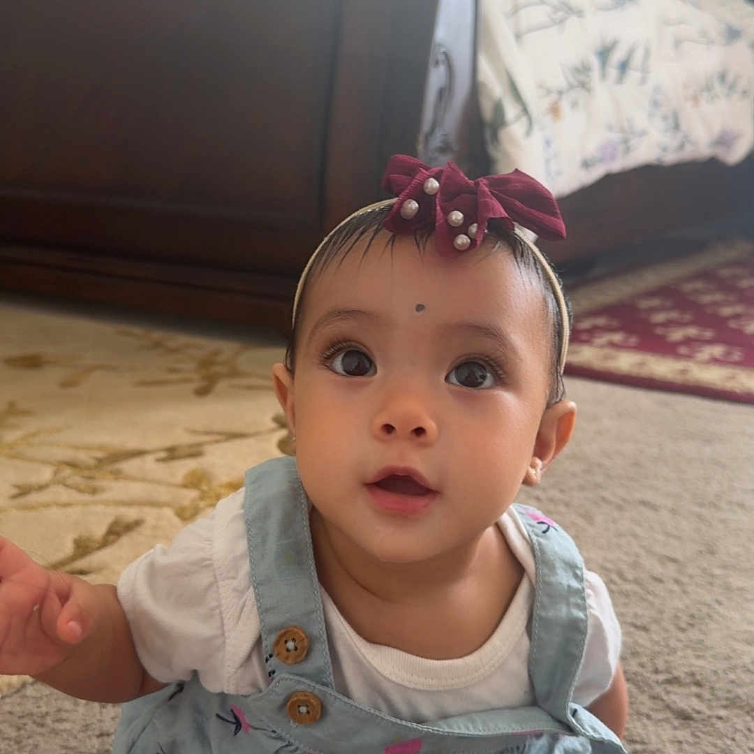 Devyani joined the competition — help win amazing prizes! baby, big_eyes, carpet, child, curious, cute, cute_baby, dress, floor, headband, home, indoor, infant, looking_up, patterned_clothing, person, portrait, sitting, white_clothing, young_child