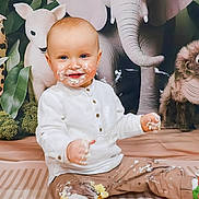 Elio participe au concours pour gagner de l'argent avec cette photo : baby, cake, messy, smiling, face, white_shirt, brown_pants, stuffed_elephant, stuffed_lamb, stuffed_monkey, floor_mat, food, cream, celebration, child, indoor, playful, cute, portrait, sitting
