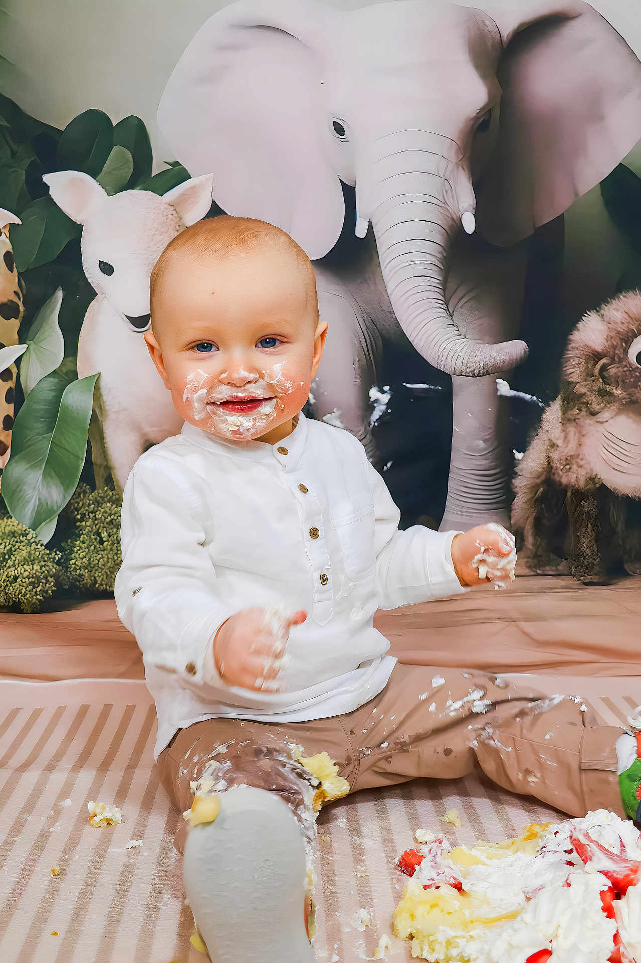 Elio participe au concours pour gagner de l'argent avec cette photo : baby, cake, messy, smiling, face, white_shirt, brown_pants, stuffed_elephant, stuffed_lamb, stuffed_monkey, floor_mat, food, cream, celebration, child, indoor, playful, cute, portrait, sitting