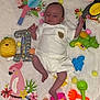 Aylan participe au concours pour gagner de l'argent avec cette photo : baby, infant, white_clothing, blanket, toys, bucket, colorful, bird_cutouts, pineapple_toy, octopus_toy, green_toy, orange_rake, yellow_pom_poms, pink_bucket, lying_down, indoors, cute, playful, smiling_toy, child