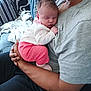 baby, sleeping, infant, person, adult, couch, pillow, blanket, clothing, pink, white, gray_shirt, black_pants, hand, arm, home_interior, relaxation, indoor, comfort, rest