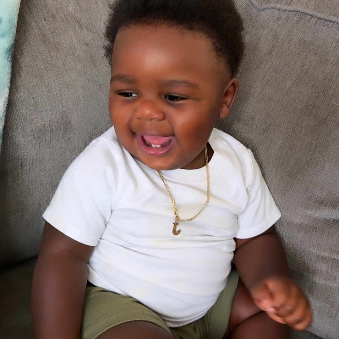 Jhream is registered to the contest to win money with this photo: baby, casual_clothing, child, couch, curly_hair, cute, dark_skin, furniture, gold_necklace, happy, home, indoor, portrait, relaxed, shorts, sitting, smiling, toddler, white_shirt, young_child