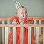 baby, child, crib, party_hat, orange_dress, pom_pom, smile, cheeks, wooden_crib, infant, cute, toddler, blue_eyes, indoor, celebration, young_child, portrait, holding_crib, happy, gown