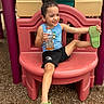 black_shorts, blue_shirt, child, colorful, curly_hair, green_shoes, happy, juice_bottle, outdoor, play_equipment, playful, playground, playground_slide, playground_surface, portrait, seated, shoe, sitting, smiling, summer