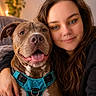 woman, dog, pet, smiling, happy, close_up, cuddle, harness, indoor, portrait, brown_dog, long_hair, casual, friendly, face, human, animal, companionship, cozy, warm_lighting