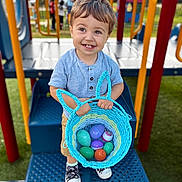Luka joined the competition — help win amazing prizes! toddler, child, playground, basket, plastic_eggs, smiling, blue_shirt, shorts, sneakers, outdoor, play_equipment, colorful, happy, toy, person, daylight, grass, metal, hand, cute